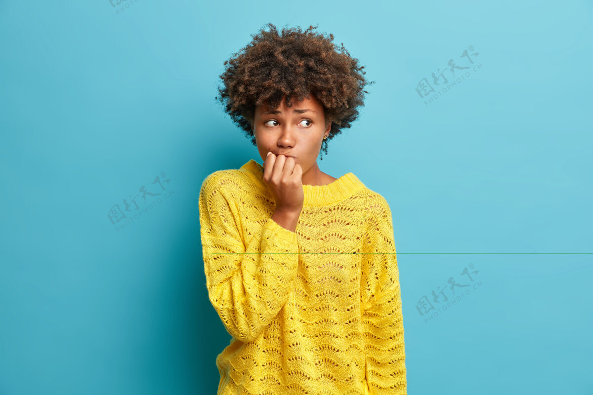 女孩紧张的女人的肖像保持手靠近嘴在重要的采访之前感到担心犹豫穿着针织黄色套头衫的东西对着蓝色的工作室墙摆姿势黑发模特问题