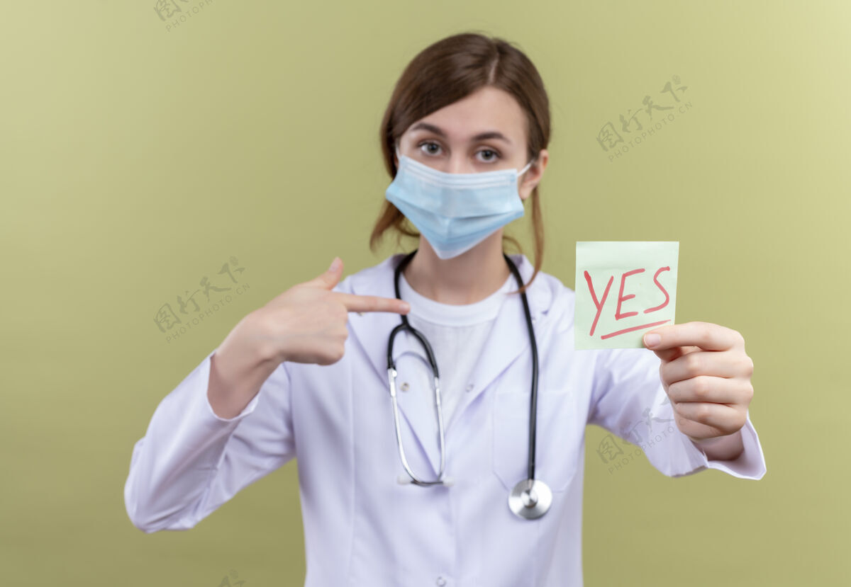 女人年轻的女医生穿着医用长袍 戴着口罩和听诊器 在隔离的绿墙上伸着“是”字条指着自己医学伸展指点