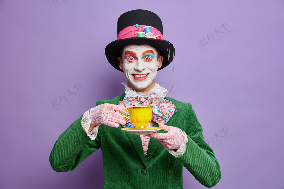 天鹅绒积极疯狂的帽匠穿着鲜艳的彩妆在派对上喝茶穿着盛装庆祝万圣节摆出快乐的姿势对着紫色的墙壁表演人物人