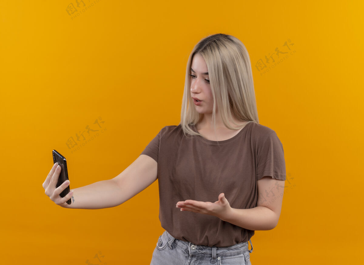 年轻年轻的金发女孩拿着手机 用手指着孤立的橙色墙上的手机指着女人手