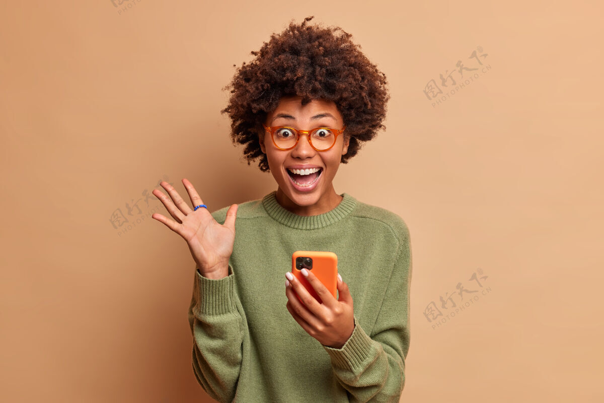 电话喜出望外的兴奋的非洲头发女人举起手掌 在收到好消息后眼睛里充满了幸福手持手机戴着眼镜和眼镜隔着棕色的墙毛衣女性技术