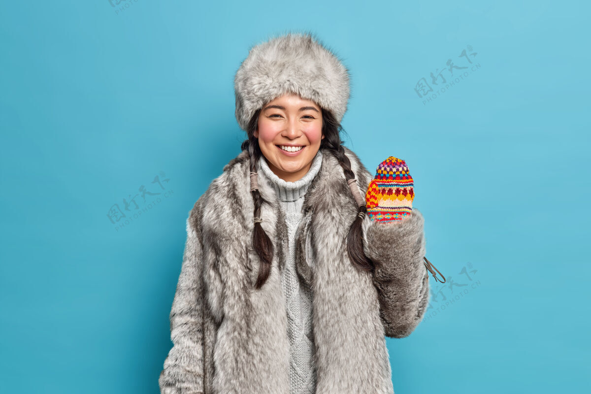 模特积极向上的黑发亚洲年轻女子身穿皮草外套 头戴帽子 手拿针织手套 微笑着愉快地享受寒假或隔着蓝色墙壁的假期北方人类黑发
