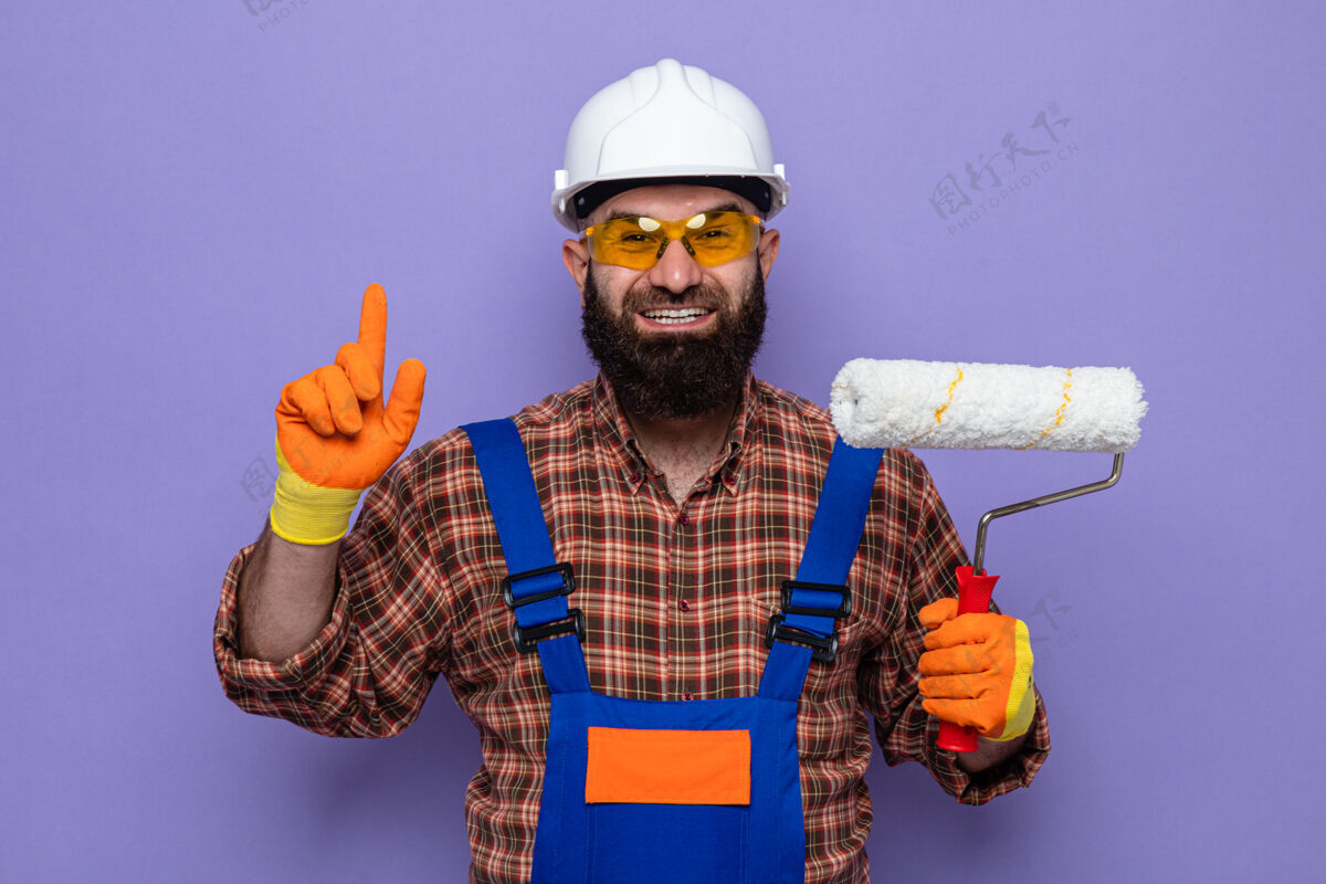 手套留着胡须的建筑工人 穿着施工制服 戴着安全帽 戴着橡胶手套 拿着油漆辊 愉快地微笑着 展示着食指有了新的想法微笑欢呼安全