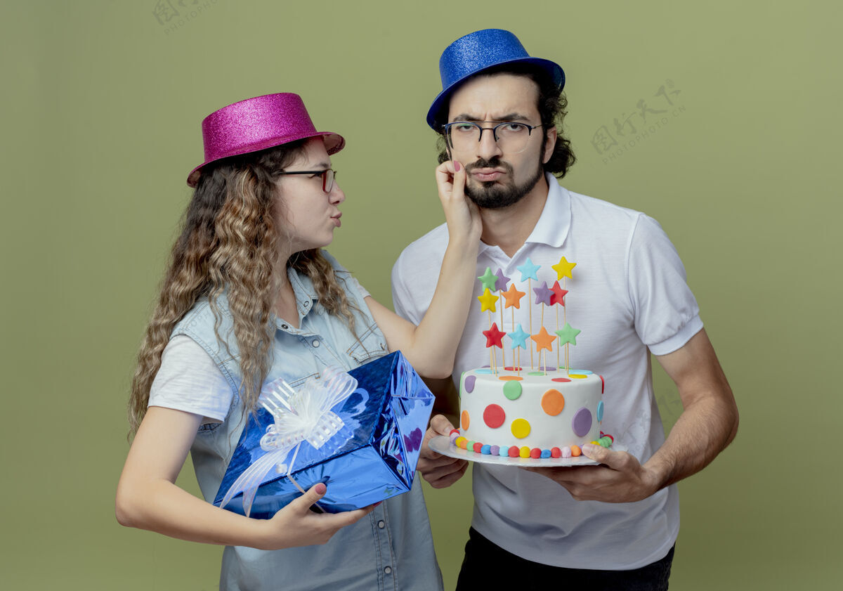 男人悲伤的年轻夫妇戴着粉色和蓝色的帽子 女孩拿着礼盒 男孩抱着脸颊 男孩拿着生日蛋糕 隔离在橄榄绿的墙上悲伤年轻蛋糕