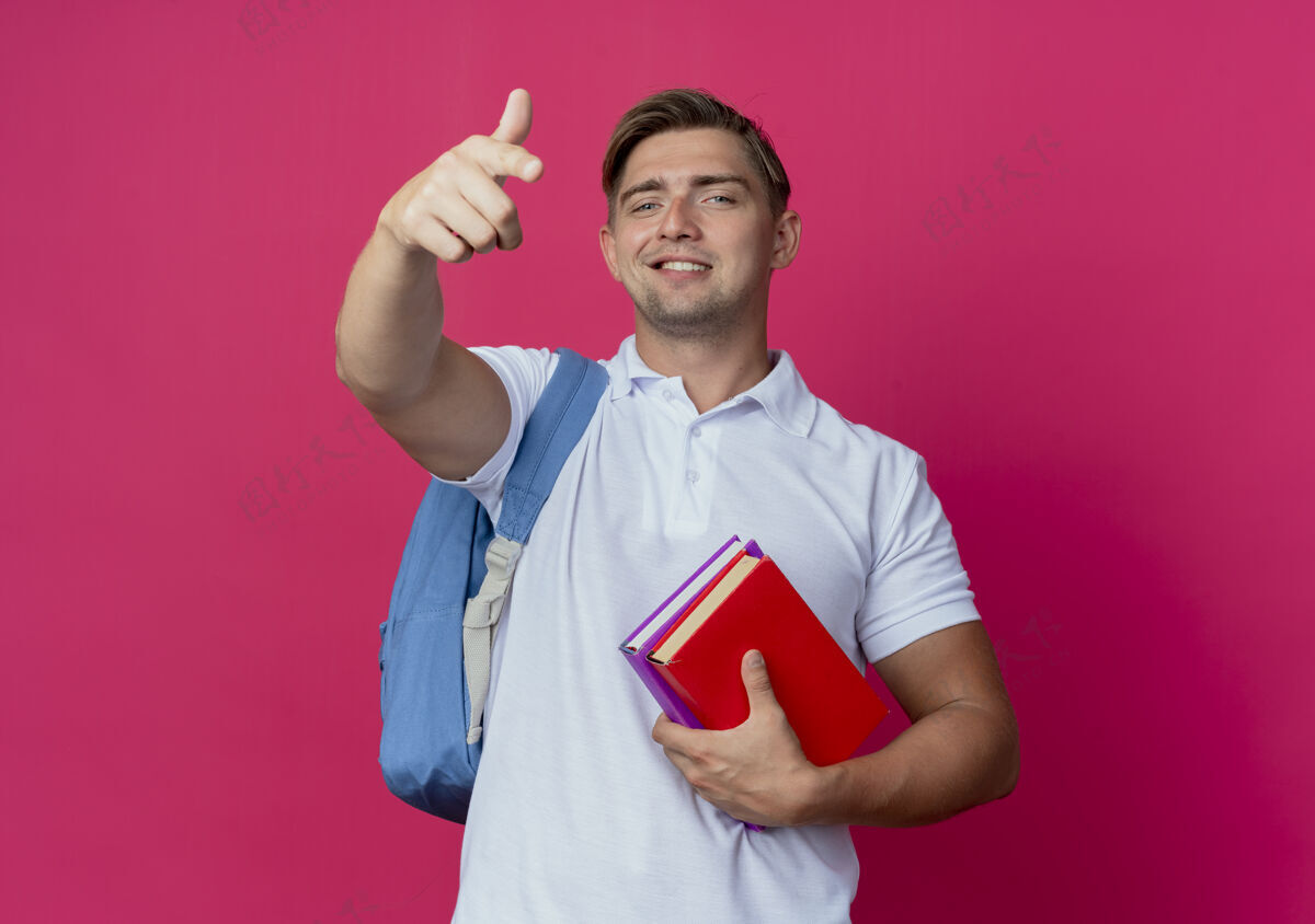 抱面带微笑的年轻英俊的男学生 背着书包 把书和点孤立在粉红色的墙上笑男学生