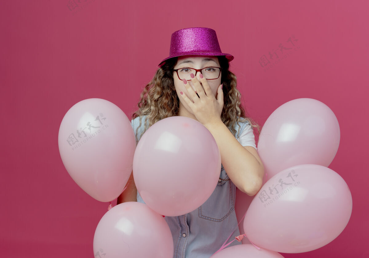 帽子被吓坏了的戴着眼镜和粉色帽子的年轻女孩站在气球中间 嘴巴被隔离在粉色的墙上戴着气球眼镜
