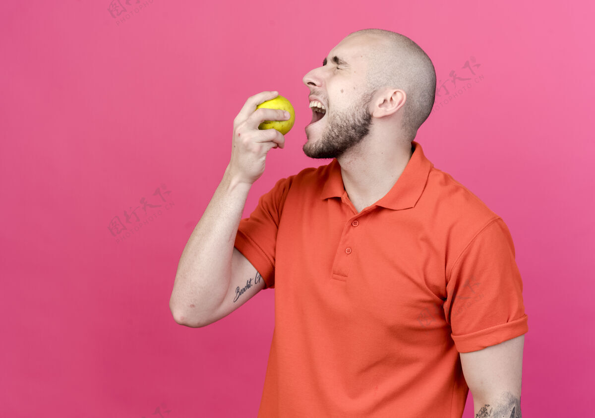 咬站在侧视图与闭着眼睛年轻的运动男子尝试咬苹果隔离在粉红色的墙壁个人资料苹果年轻