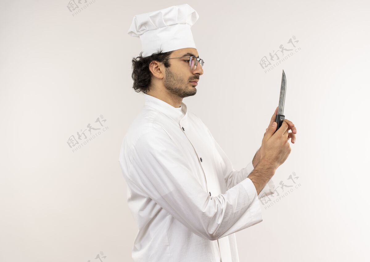 观点站在剖面图年轻的男厨师穿着厨师制服 戴着眼镜 拿着刀看着隔离在白色墙上的墙立场厨师穿