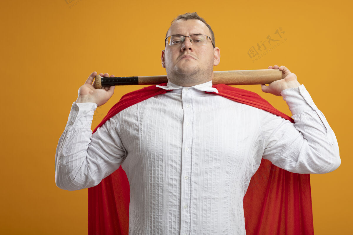 成人自信的成年斯拉夫超级英雄 穿着红色斗篷 戴着眼镜 脖子后面拿着棒球棒 隔离在橙色的墙上橙色眼镜抱