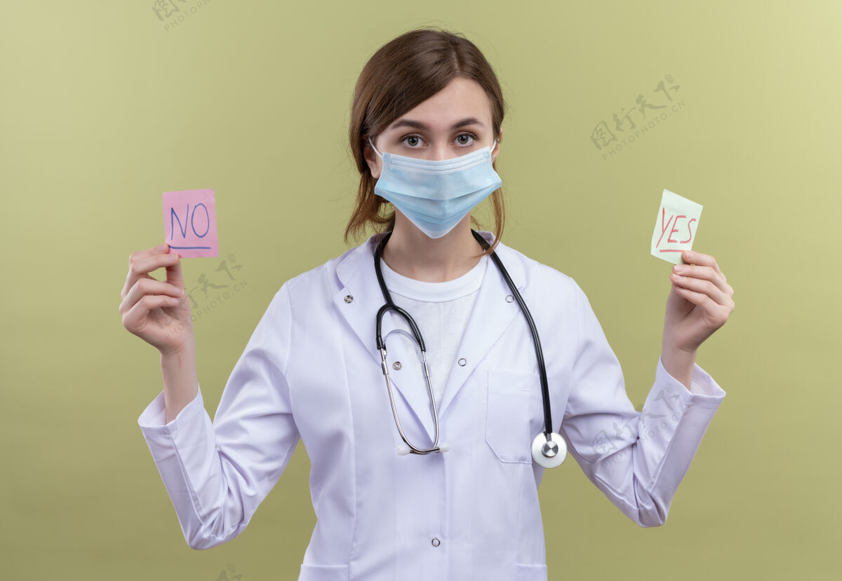 年轻年轻的女医生穿着医用长袍 戴着口罩和听诊器 拿着是和否的纸条 看着隔离的绿色墙壁抱着医生长袍
