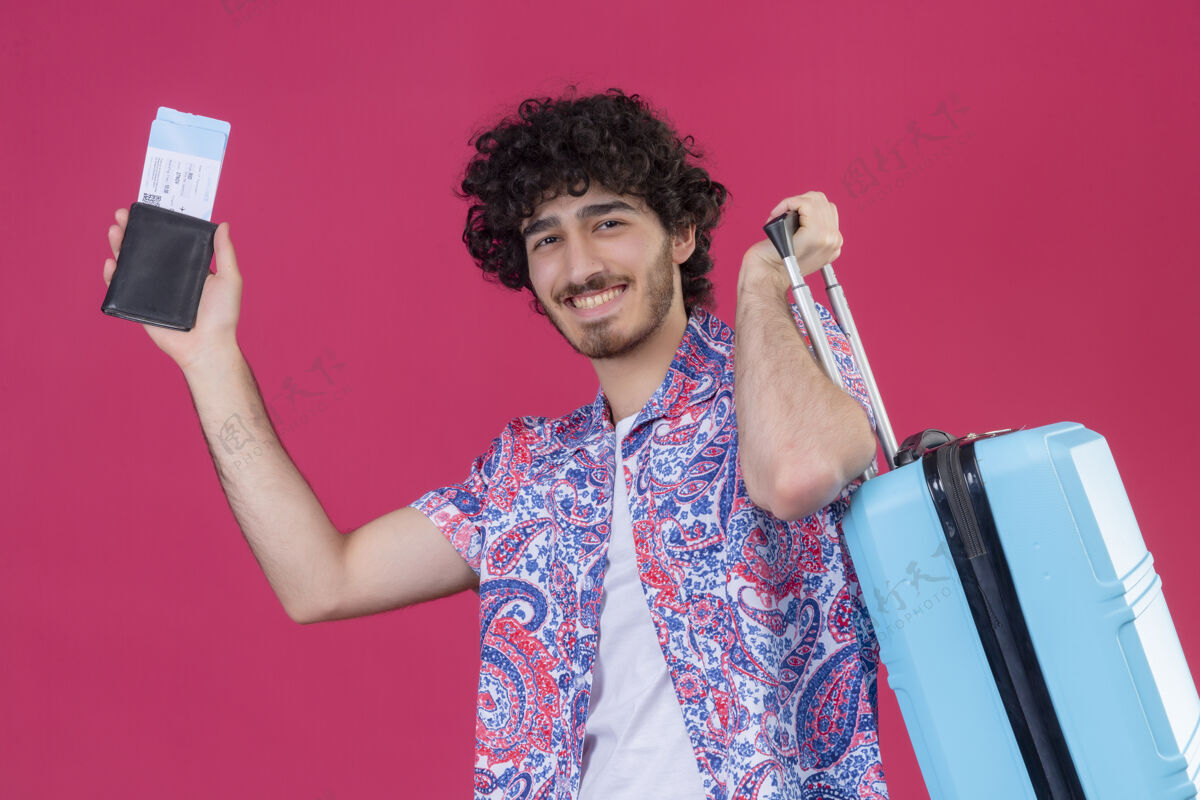 男人微笑着的年轻英俊的卷发旅行者 手里拿着钱包 机票和手提箱 孤零零地站在粉红色的墙上钱包卷发帅哥