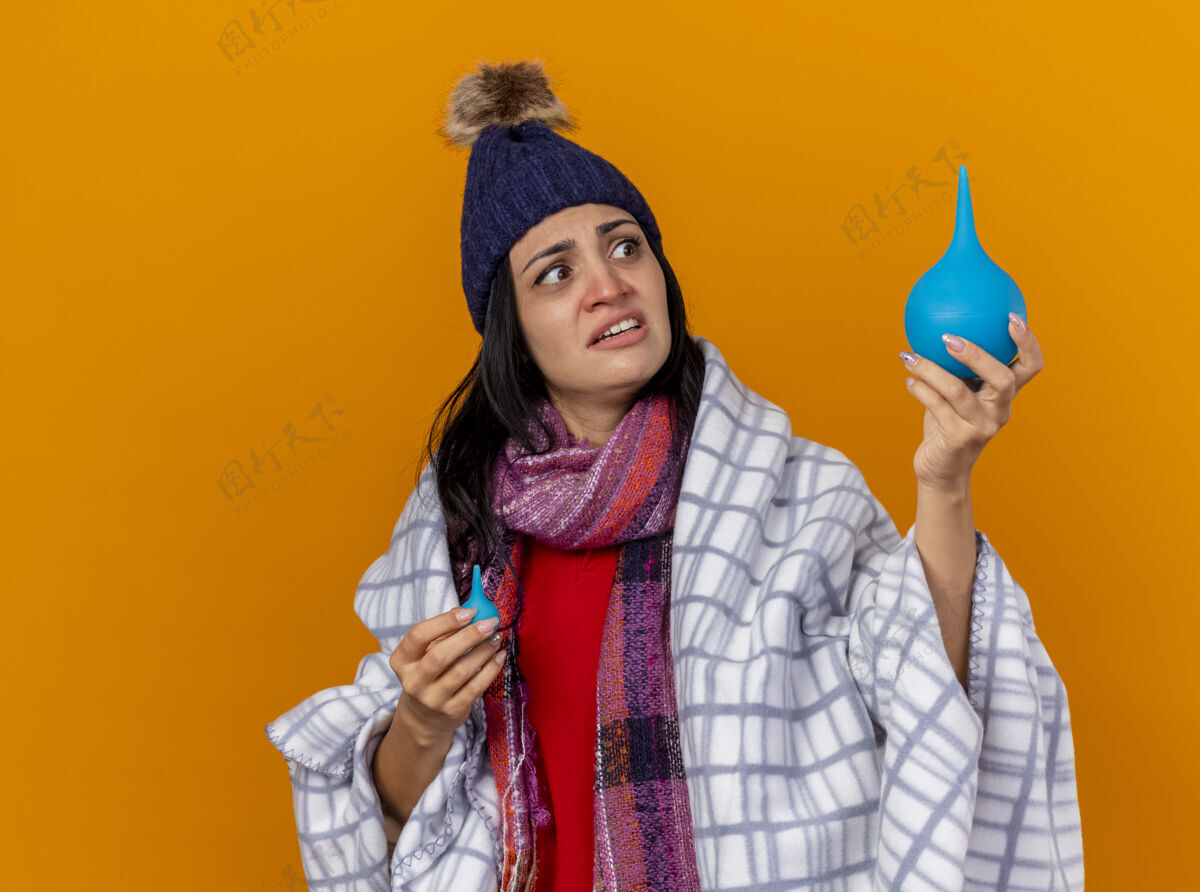 灌肠印象深刻的年轻白种人生病的女孩戴着冬天的帽子和围巾包裹在格子布拿着灌肠看着一个大的橙色墙上孤立的复制空间印象包裹橙色
