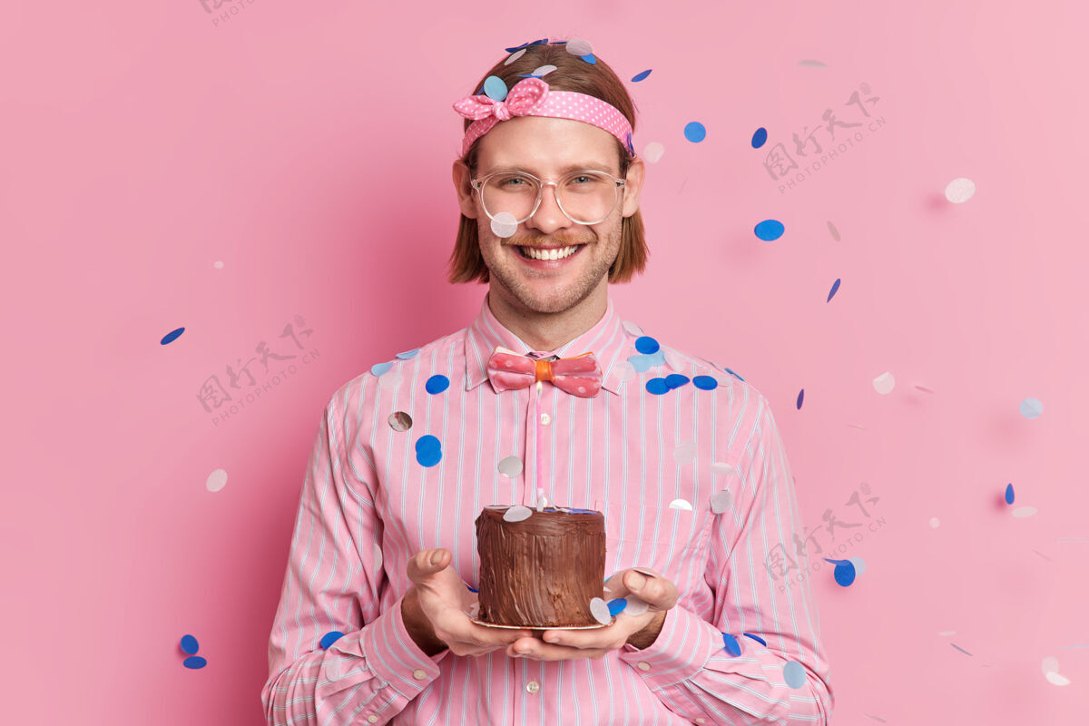成人快乐的成年男子庆祝在公司工作一年拿着小蛋糕庆祝同事们的祝贺微笑着高兴地戴着头带条纹衬衫和蝴蝶结科菲蒂落在他身上快乐生日五彩纸屑