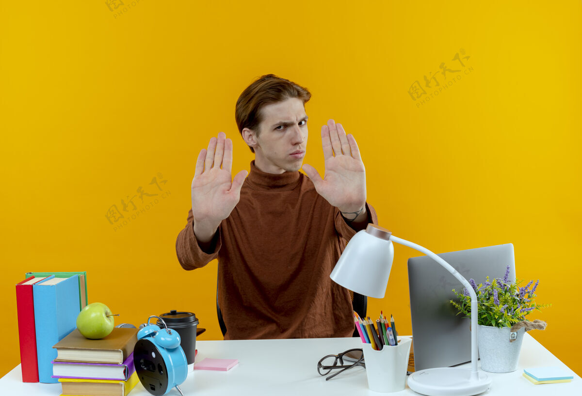 男孩严格的年轻学生男孩坐在书桌旁 在黄色的墙上显示停止手势工具学生坐着
