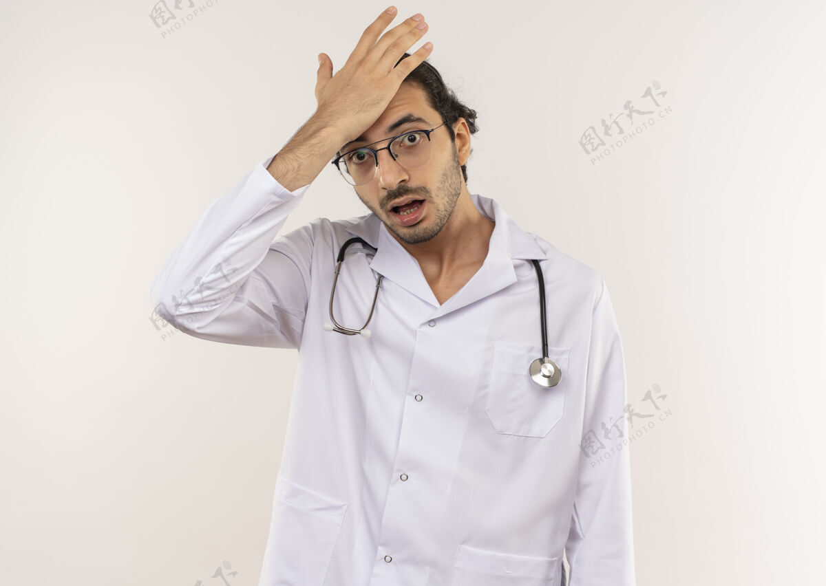 戴戴眼镜的年轻男医生穿着白色长袍 手持听诊器 将手放在额头上 在隔离的白色墙壁上留有复印空间眼镜听诊器男性