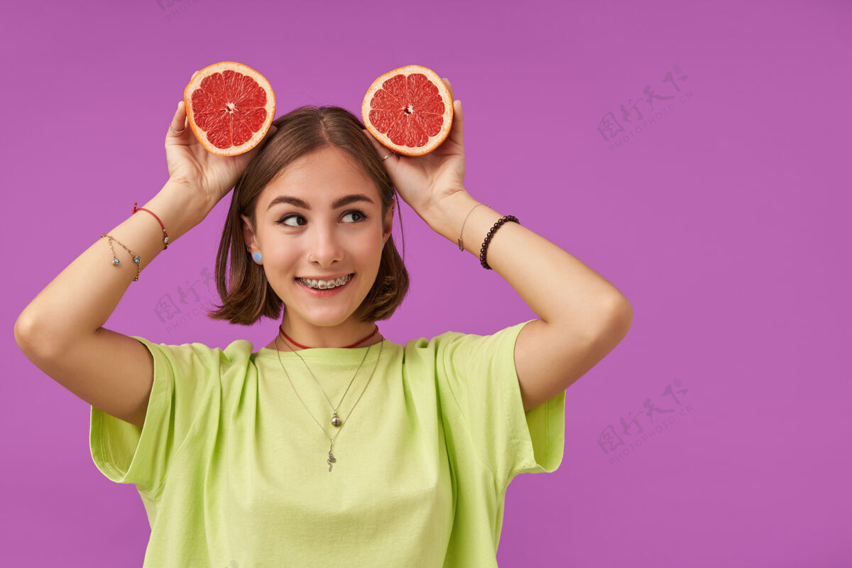 独自漂亮的女人 深色短发 看着右边紫色墙上的复制空间 头上戴着葡萄柚 穿着绿色t恤 戴着牙套 手镯和项链穿孔项链牙齿