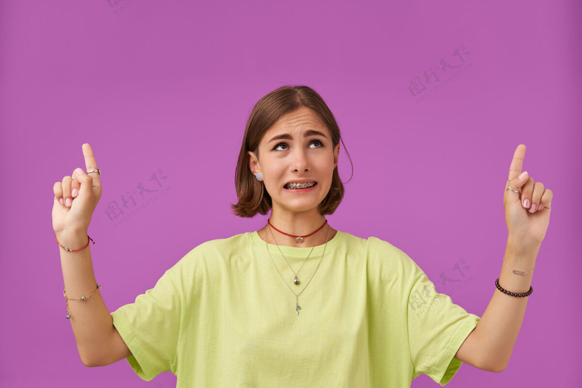 青少年女学生 年轻不快乐的女士 深色短发手指指向紫色墙上的复印空间穿着绿色t恤 牙套 项链 手镯和戒指短发女孩女士