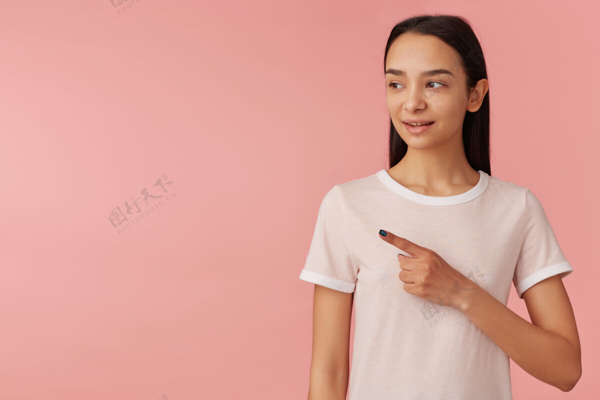 指向可爱的女人 长着黑色长发的漂亮女孩穿着白色t恤人们的概念看着并用食指指着复制空间的左侧 隔离在粉彩粉色的墙上单独女人学生