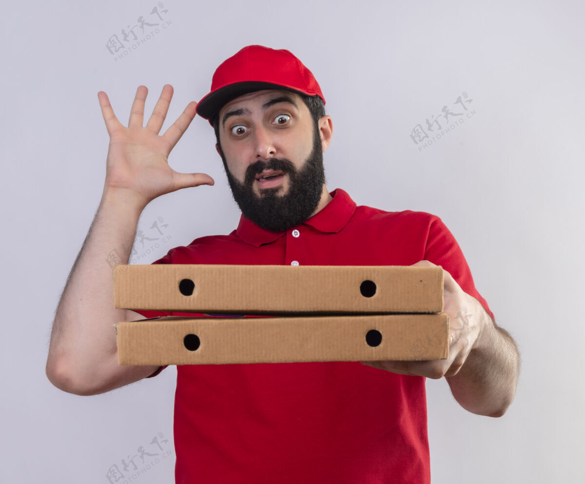 帅气令人印象深刻的年轻英俊的白人送货员穿着红色制服 戴着帽子 看着披萨盒举起手孤立在白色盒子披萨年轻