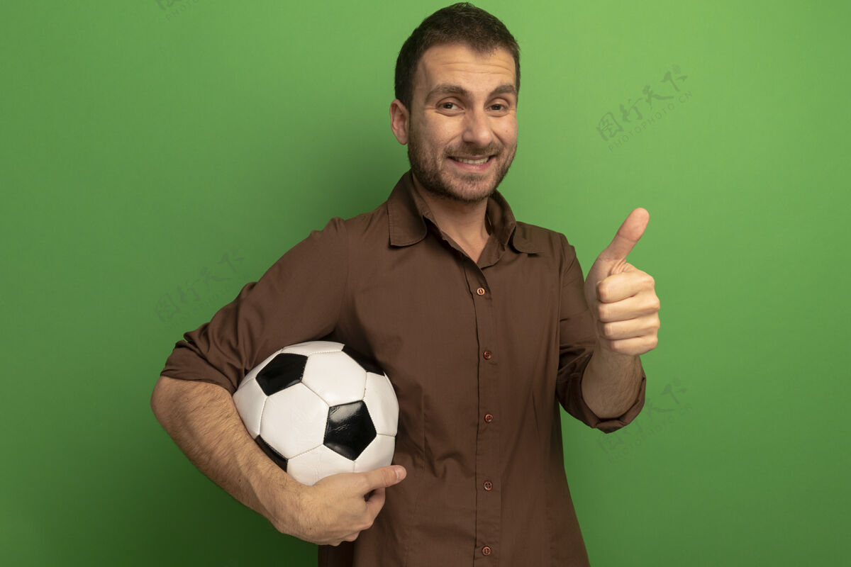 握着微笑的白人年轻人拿着足球看着相机 在绿色的背景上 用复制空间孤立地竖起大拇指相机微笑年轻人