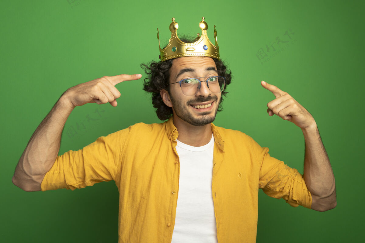 眼镜微笑着的年轻英俊的白人男子戴着眼镜和王冠指着自己的王冠看着隔离在绿色背景下的相机微笑年轻帅气
