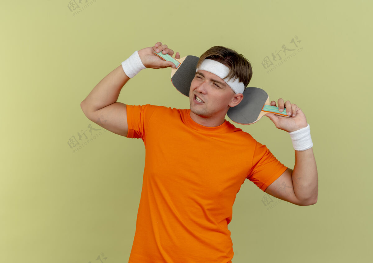 乒乓球自信的年轻帅气的运动型男人戴着头带和腕带 在脑后拿着乒乓球拍 看着橄榄绿上孤立的一面头带腕带穿
