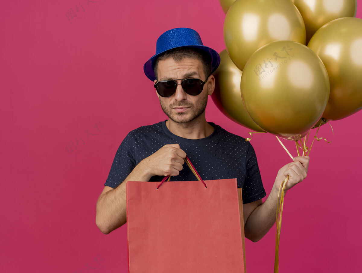 帽子严肃英俊的白种人 戴着太阳眼镜 戴着蓝色的派对帽 手里拿着氦气球和纸购物袋 在粉色背景上隔离开来 还有复印空间聚会帅气包