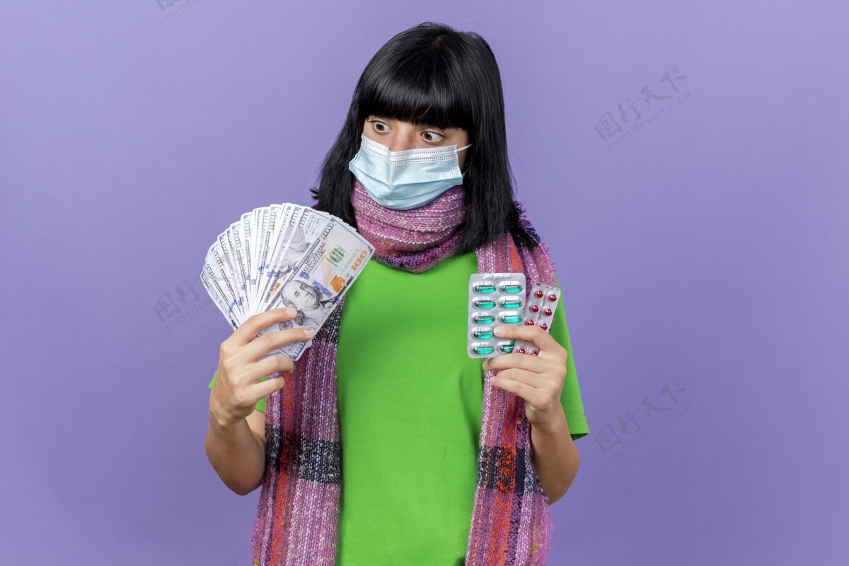 年轻印象深刻的年轻病高加索女孩戴着面具和围巾拿着钱和胶囊包看着钱孤立的紫色背景与复制空间胶囊围巾女孩