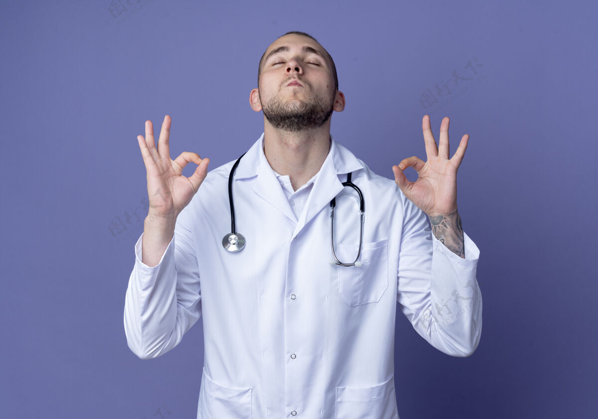 标志自信的年轻男性医生穿着医用长袍和听诊器 闭着眼睛做着正常的体征 隔离在紫色的复印空间里长袍佩戴男性