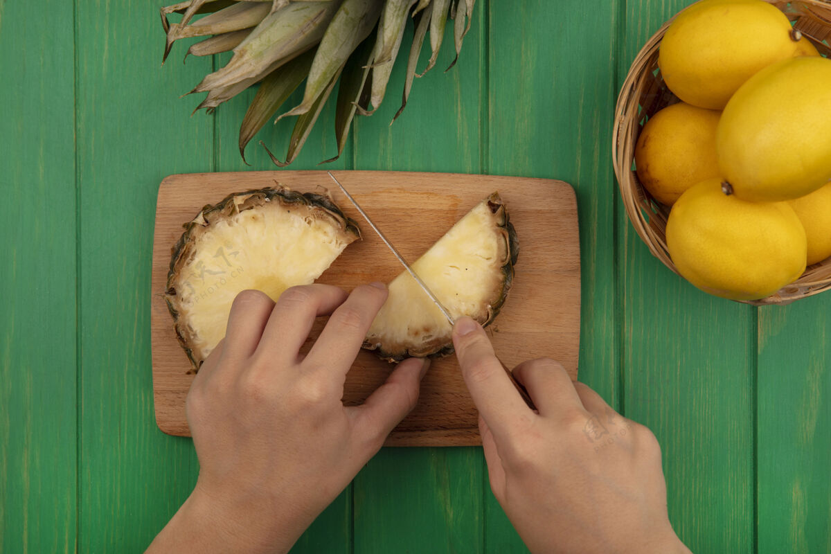 菠萝俯视图中的女性手切甜菠萝在一个木制的厨房板与刀子柠檬桶上的绿色木墙上切割新鲜手