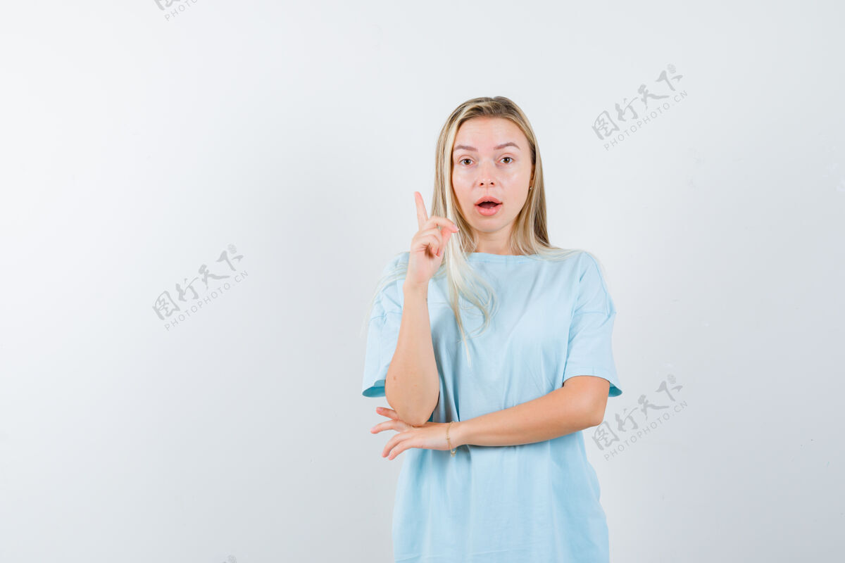 提高金发女孩举起食指在尤里卡的手势 同时在手肘下牵着手在蓝色t恤 看起来很理智 前视图肘部健康旅游