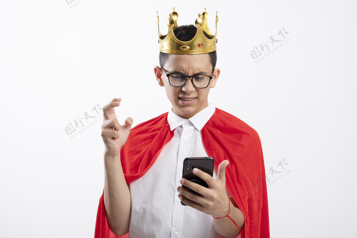 年轻穿着红斗篷戴着眼镜戴着王冠的年轻超级英雄男孩看着手机 手伸进空气中 白色背景上留着空白超级英雄手机电话