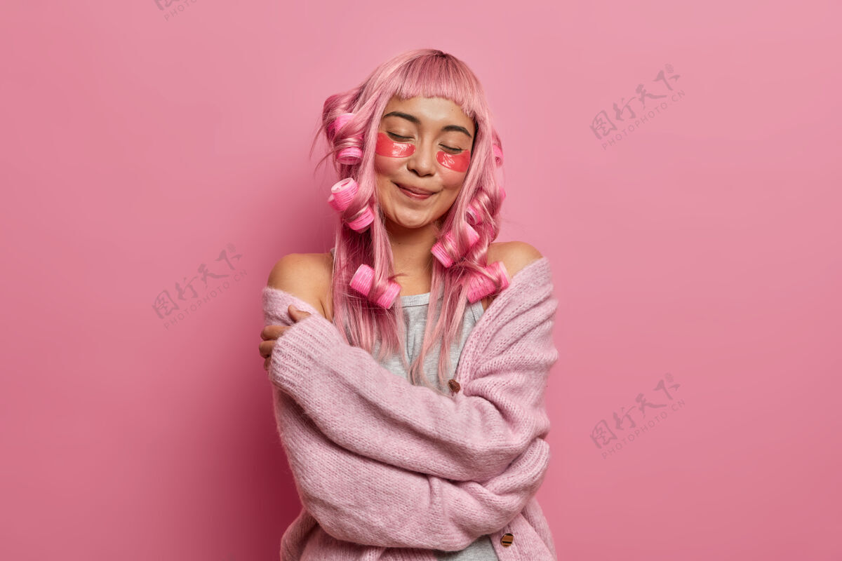 美容快乐的微笑迷人的女士闭上眼睛 拥抱自己 感觉舒适的新针织套头衫 应用美容垫和卷发长粉红色的头发漂亮穿姿势