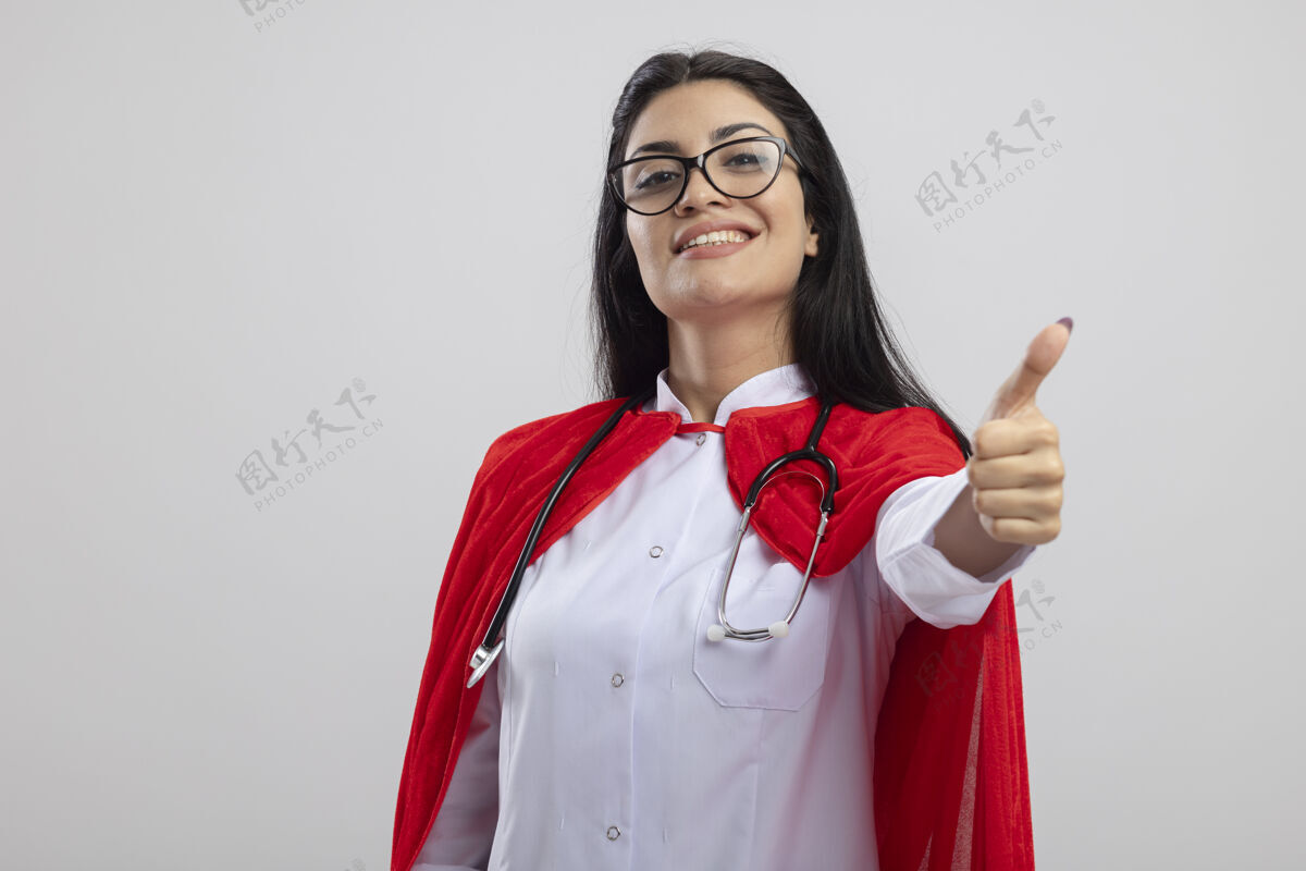 听诊器带着微笑的年轻白人超级英雄女孩戴着眼镜和听诊器看着相机 在白色背景上孤立地竖起大拇指 留有复印空间年轻复制穿