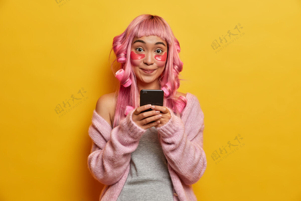 完美微笑积极的亚洲女人手里拿着手机 查看邮箱 神采奕奕 有着粉红色的头发 戴着滚轴检查纯洁黄色