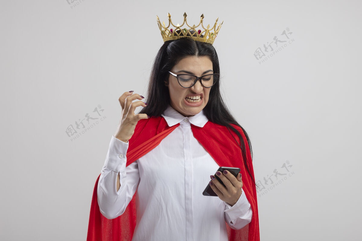 佩戴愤怒的年轻白种人超级英雄女孩戴着眼镜 戴着王冠 手举着手机看着 在白色的背景上与外界隔绝 留着复印空间年轻手愤怒