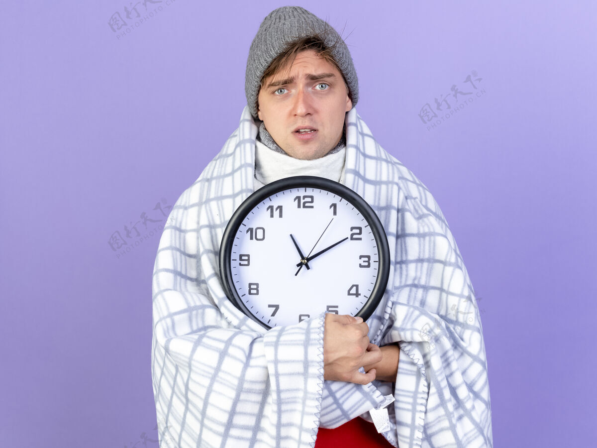 冬天印象深刻的年轻英俊的金发病男子戴着冬天的帽子和围巾包裹在格子布拿着时钟看着相机在紫色背景与复制空间隔离紫色空间年轻