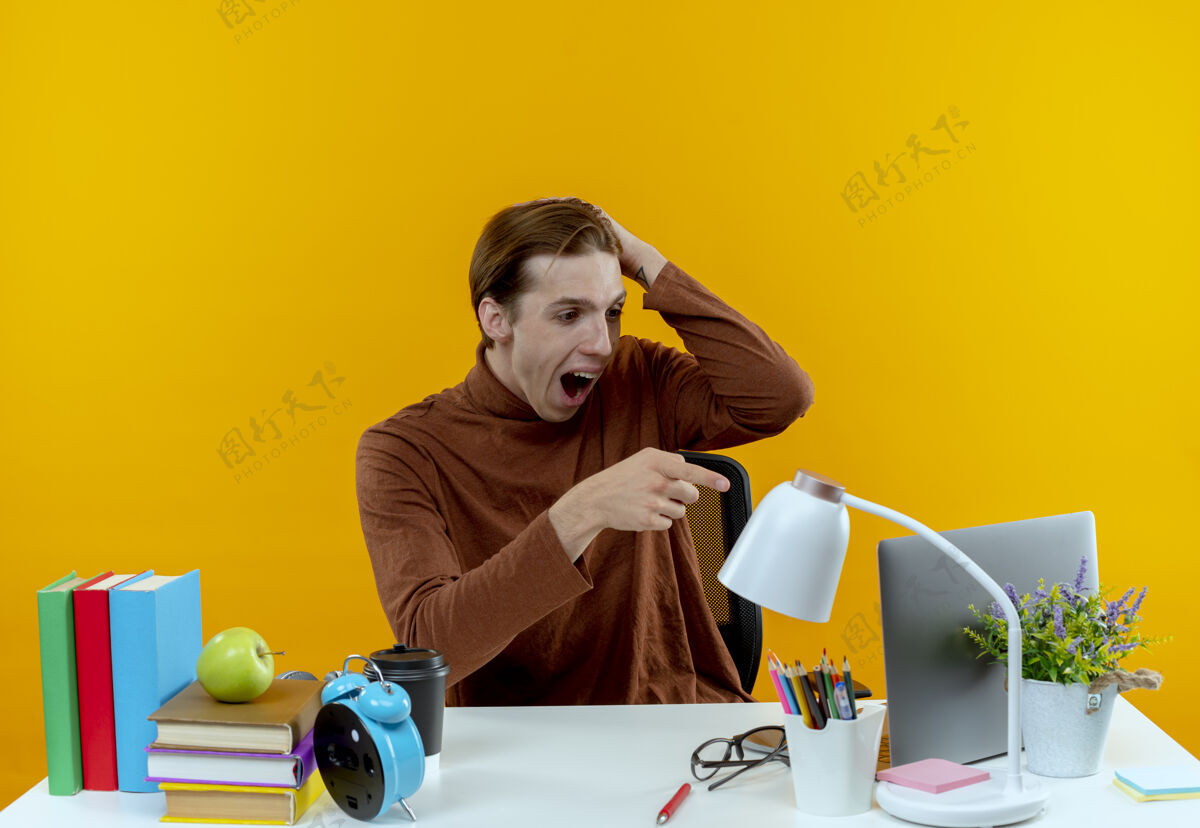 黄色被吓坏了的小男孩坐在书桌旁 手里拿着学习工具 指着笔记本电脑 把手放在黄色的头上桌子笔记本电脑坐着