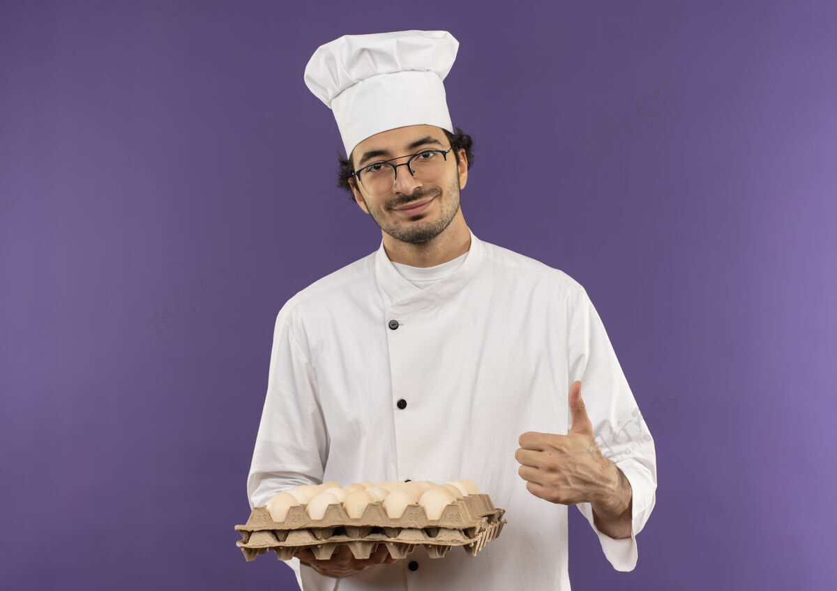 烹饪高兴的年轻男厨师穿着厨师制服 戴着眼镜 手里拿着一批鸡蛋 大拇指朝上放在紫色上制服鸡蛋紫色