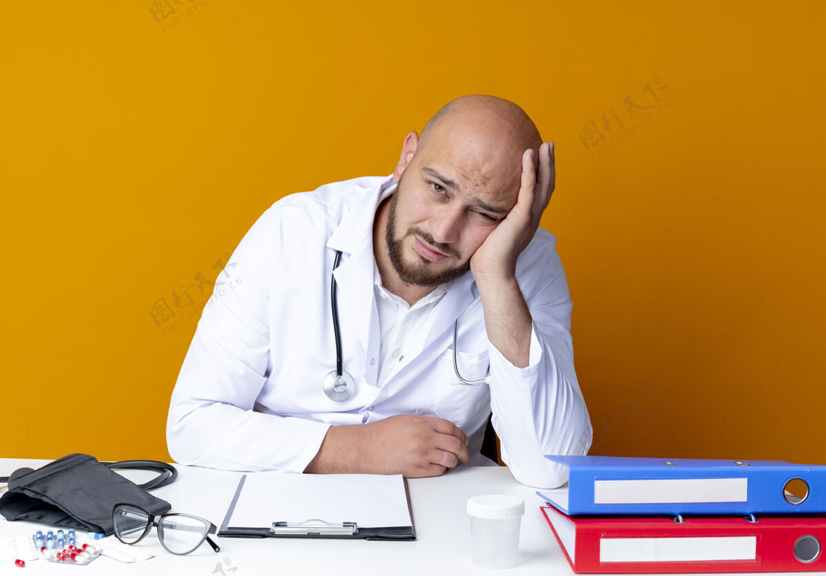 医疗疲惫的年轻秃头男医生穿着医用长袍和听诊器坐在办公桌旁 手放在橘子色的脸颊上疲倦医生工作