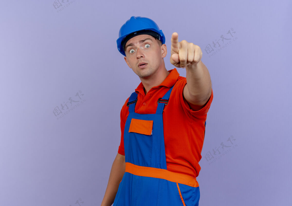 年轻身穿制服和安全帽的年轻男性建筑工人惊讶地指着紫色的摄像机头盔安全点