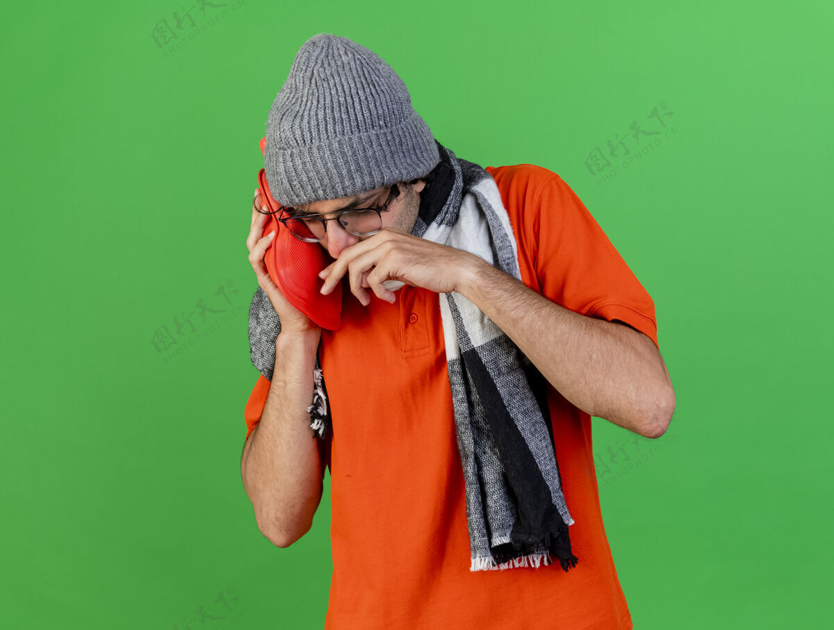 公民愁眉不展的年轻病人戴着眼镜 戴着冬天的帽子和围巾把热水袋放在脸上擦鼻子用手隔离在绿色的墙上围巾水热
