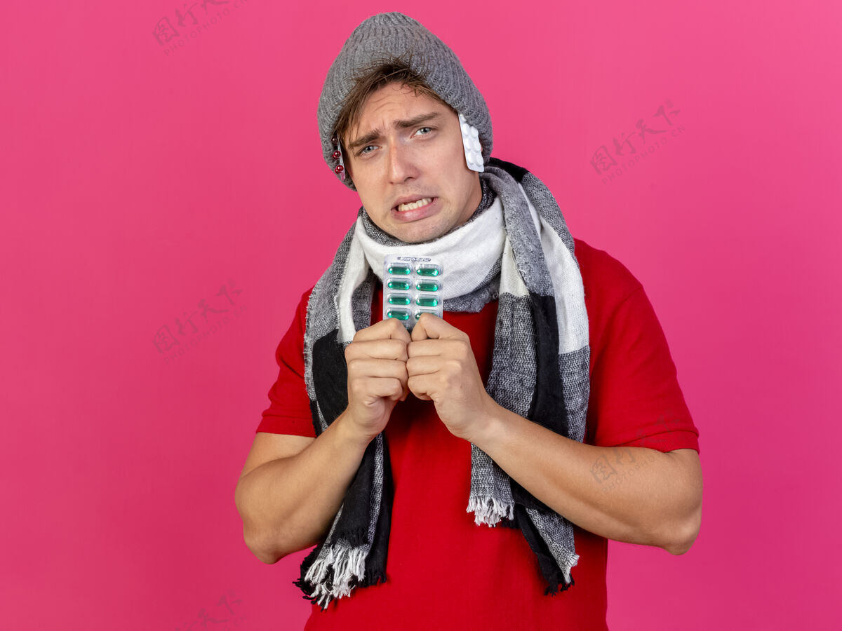 年轻人愁眉不展的年轻英俊的金发病男子戴着冬天的帽子和围巾拿着一包药丸一包一包的药丸在帽子下面看着隔离在粉红色墙上的前面疾病人市民