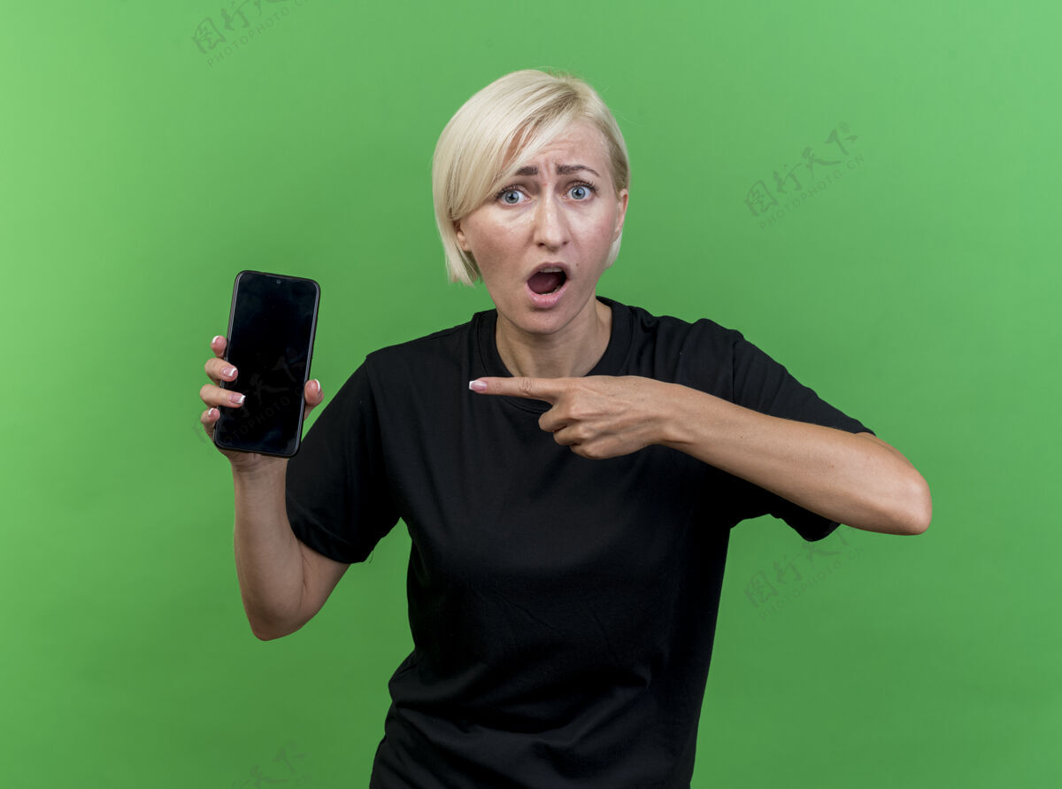 人印象深刻的中年金发女人看着前面展示的手机指着它隔离在绿色的墙上印象脸人