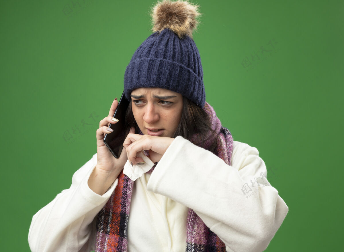 壁板愁眉不展的年轻病女人穿着长袍 戴着冬天的帽子和围巾 讲着电话 拿着餐巾 手放在嘴边 看着隔离在绿色墙上的一面帽子手电话