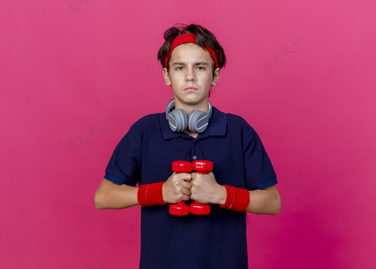 戴自信的年轻帅气的运动男孩戴着头带和腕带 脖子上戴着耳机 戴着牙套 举着哑铃 看着深红的墙上孤立的前方表情人运动