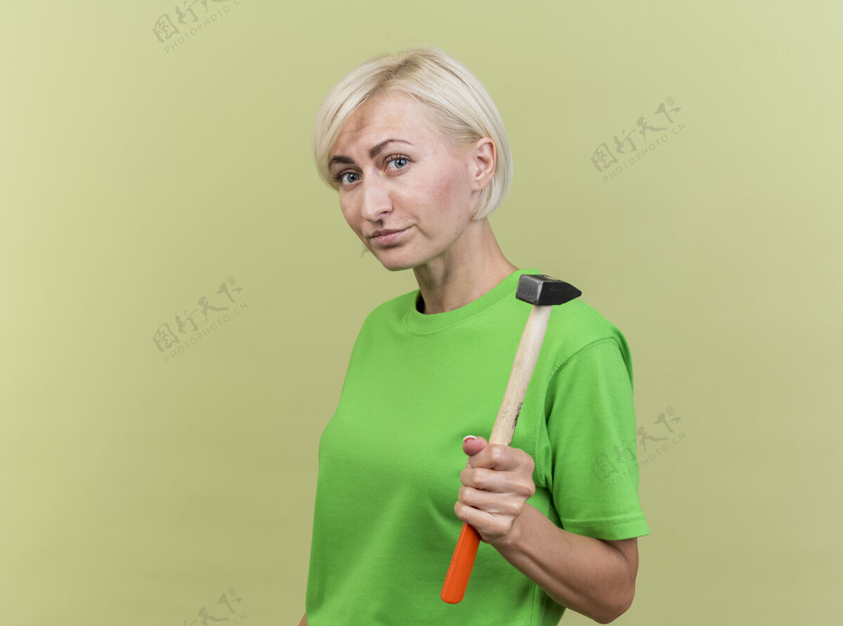女人自信的中年金发女人站在侧视图中 看着橄榄绿墙上孤立的手锤衣服金发自信