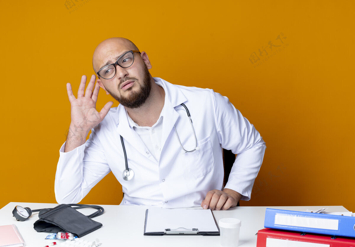 手势想着年轻的秃头男医生穿着医用长袍 戴着听诊器坐在眼镜里医生听诊器坐着