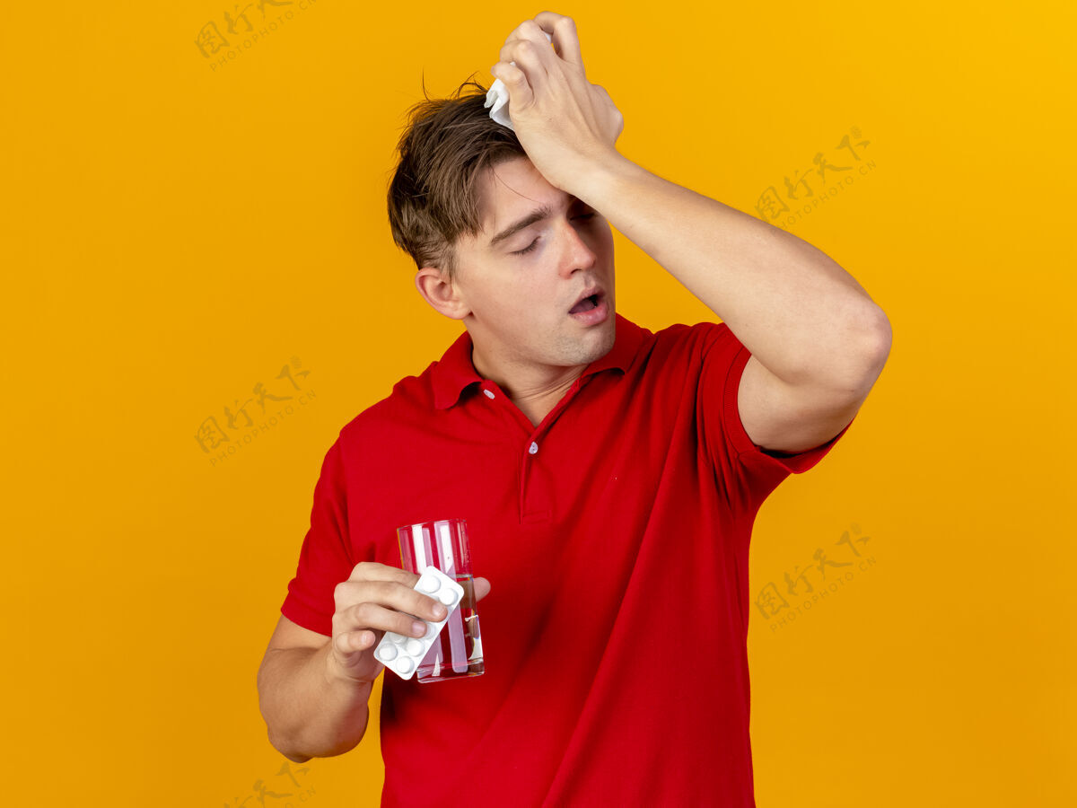 餐巾纸疼痛的年轻英俊的金发病患拿着一包药片一杯水和餐巾摸着头 闭着眼睛在橘色的墙上孤立着头痛药片触摸人