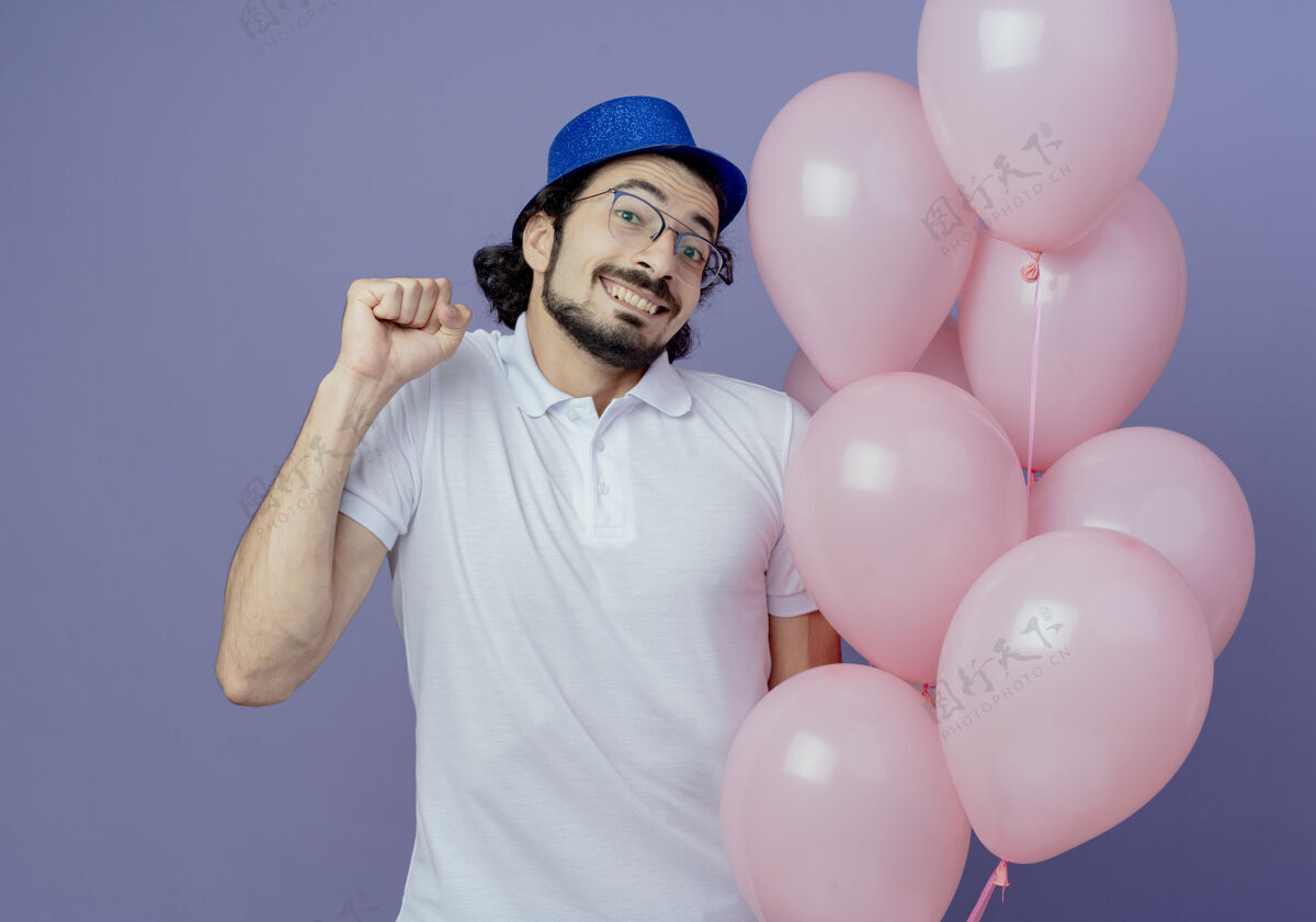 帽子笑容可掬的帅哥戴着眼镜 戴着蓝色的帽子 手里拿着气球 摆着“是”的手势 孤立地站在紫色上帅气戴着是的